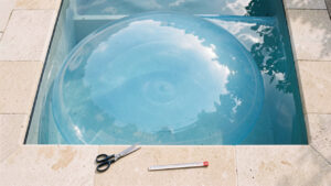 Vue de dessus d’une piscine rectangulaire recouverte d’une bâche à bulles bleue, avec un mètre ruban et des ciseaux posés sur la margelle, éclairée par une lumière naturelle.