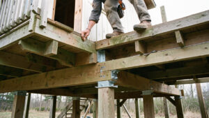 Un homme renforce le plancher en bois d’une cabane sur pilotis à l’aide de poutres et de supports métalliques.